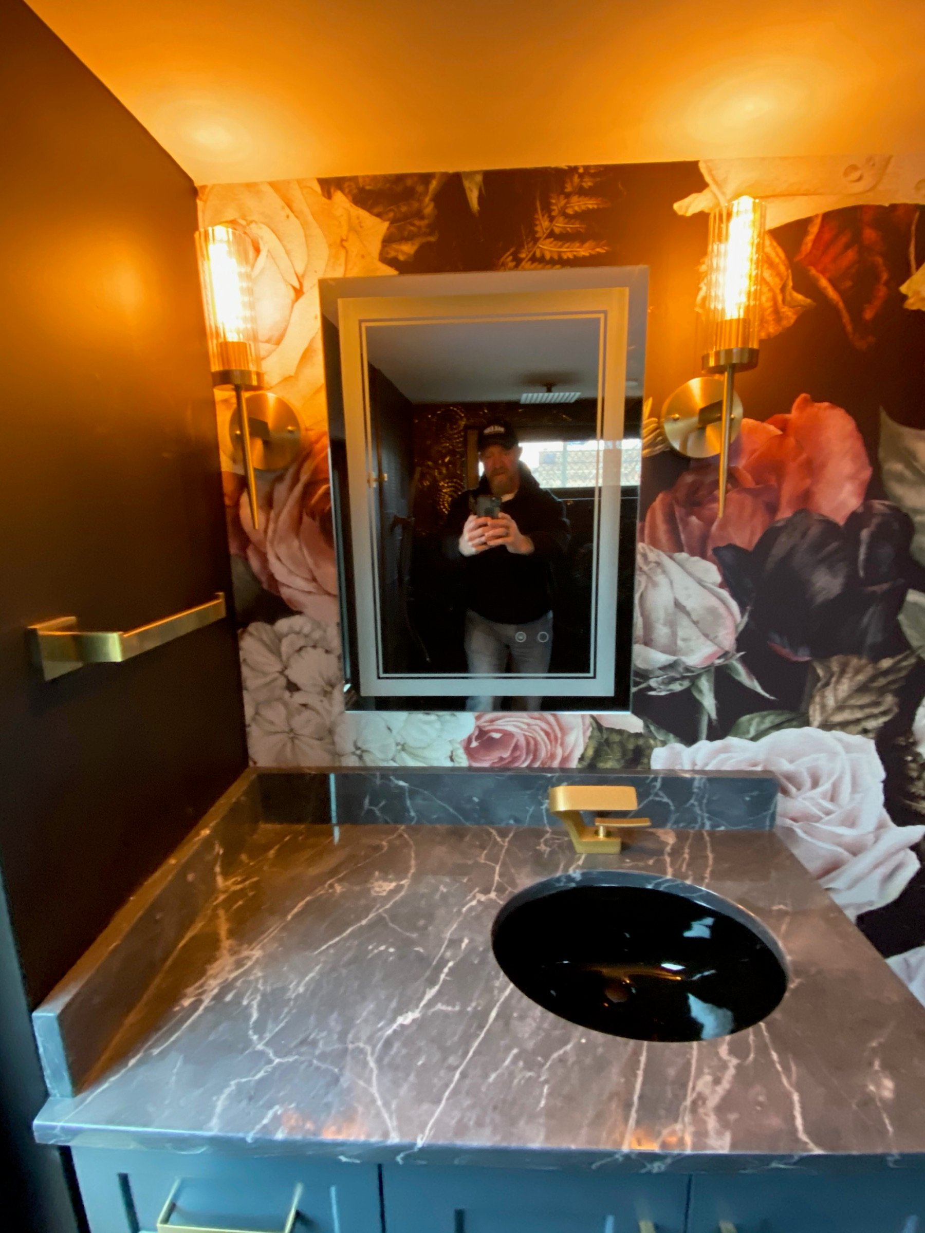 Compact bath vanity with floral wallcovering, dark stone top, and brass lighting